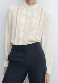 Beige blouse met een gerimpelde kraag, verticale streepjes en kantaccenten. Lange mouwen met knoopmanchetten. Draagt met donkere, op maat gemaakte broeken.