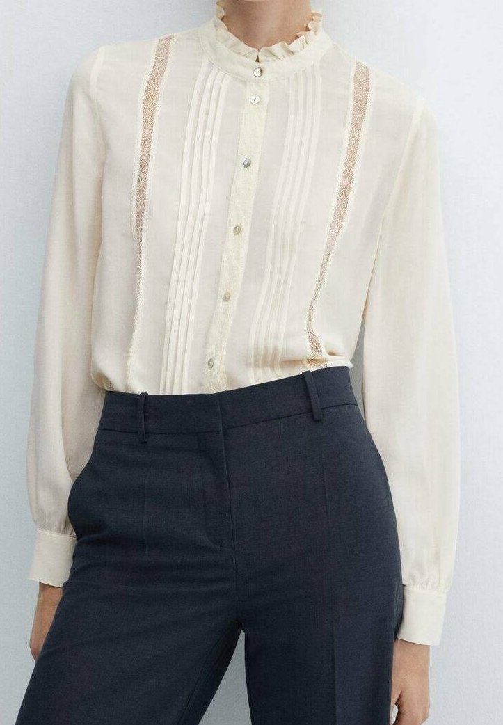 Beige blouse met een gerimpelde kraag, verticale streepjes en kantaccenten. Lange mouwen met knoopmanchetten. Draagt met donkere, op maat gemaakte broeken.