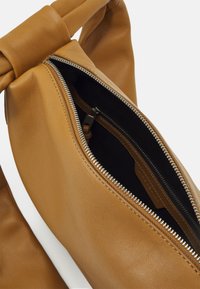Sac en cuir tan avec une anse en forme de nœud, doté d'une fermeture éclair argentée et d'un intérieur sombre. Le sac a une texture douce et un design minimaliste.