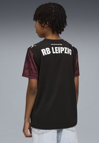 Czarna koszulka sportowa z krótkim rękawem. Rękawy ozdobione są czerwonym wzorem geometrycznym. Z tyłu widnieje napis "RB LEIPZIG" dużymi białymi literami.