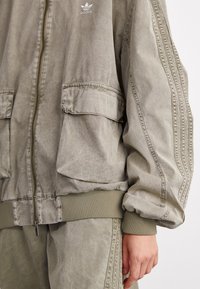 Olive green bomber jacket made of soft, textured fabric. Features elastic cuffs, a zip closure, and two front pockets. adidas logo on chest.