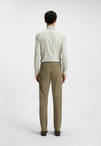 Chemise boutonnée beige clair à manches longues, associée à un pantalon vert olive avec des poches arrière. La tenue est complétée par des chaussures sombres.