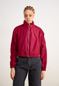 Red cropped jacket with a high collar, zipper closure, elastic cuffs, and a textured finish. Features a small logo on the chest.