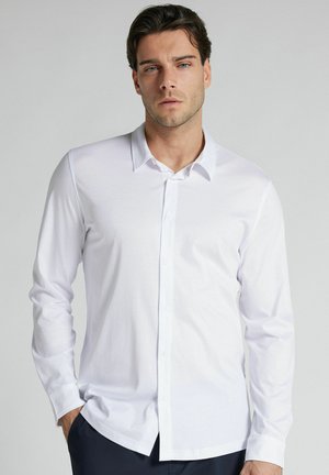 Mand med mørkt hår iført en hvid skjorte med lange ærmer og knapper samt mørke bukser, står foran en ensfarvet lys grå baggrund.