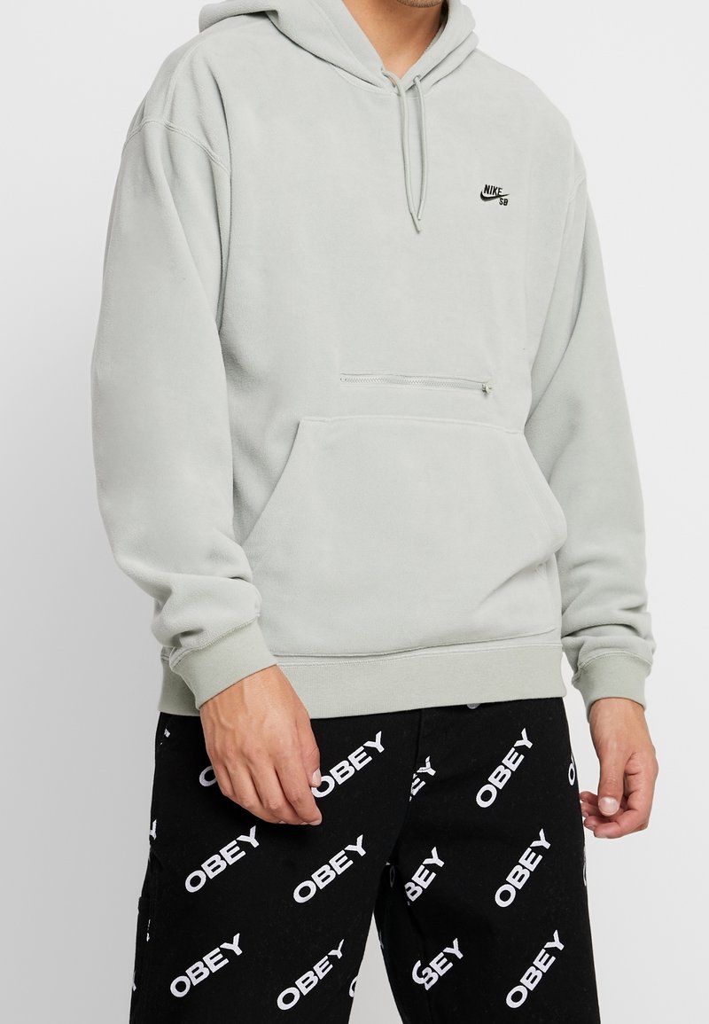 Sweat à capuche gris clair à finition texturée, capuche ajustable, poche kangourou et petit logo noir sur la poitrine. Associé à un pantalon noir à motif.