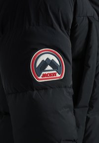 Manche de veste matelassée noire avec un patch circulaire montrant des sommets de montagne stylisés et le mot « JACK1T » en lettres rouges.