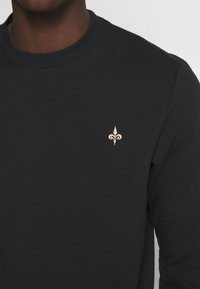 Schwarzer Baumwoll-Hoodie mit weicher Textur und Rundhalsausschnitt. Enthält ein goldbesticktes Fleur-de-Lis-Detail auf der Brust.