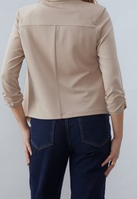 Béžová, lehká bunda s 3/4 rukávy, jednoduchý design a šev na zadní straně; doplněná tmavě modrými džíny s zadními kapsami.