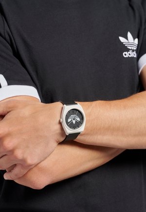 Digitalni sat s crnim teksturiranim remenom, kvadratnim srebrnim kućištem i crnim brojčanikom koji sadrži bijele oznake sati i dizajn logotipa.