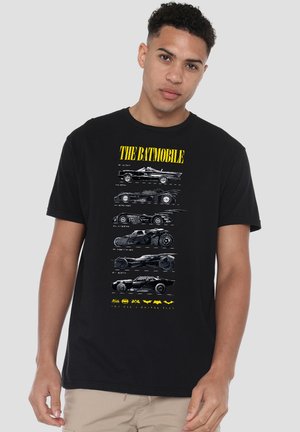 Junger Mann trägt ein schwarzes T-Shirt mit sechs verschiedenen weißen Batmobile-Autodesigns und gelbem Text "THE BATMOBILE" darüber.
