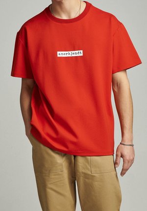 Mies, joka pukeutuu punaisiin lyhythihaisiin T-paitoihin, joissa on musta "anerkjendt" -teksti valkoisella suorakaiteella. Hän on yhdistänyt sen beigeihin housuihin ja oikean ranteen hopeaketjun rannekoruun.