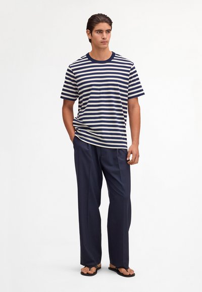 Giovane uomo in piedi che indossa una maglietta a maniche corte a righe blu navy e bianche, pantaloni larghi blu navy e infradito nere su sfondo bianco.