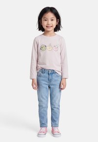 Ένα παιδί που χαμογελά φοράει ροζ μπλούζα με μακριά μανίκια και τρία παιχνιδιάρικα σχέδια ντόνατ, ανοιχτό μπλε τζιν και ροζ αθλητικά παπούτσια.