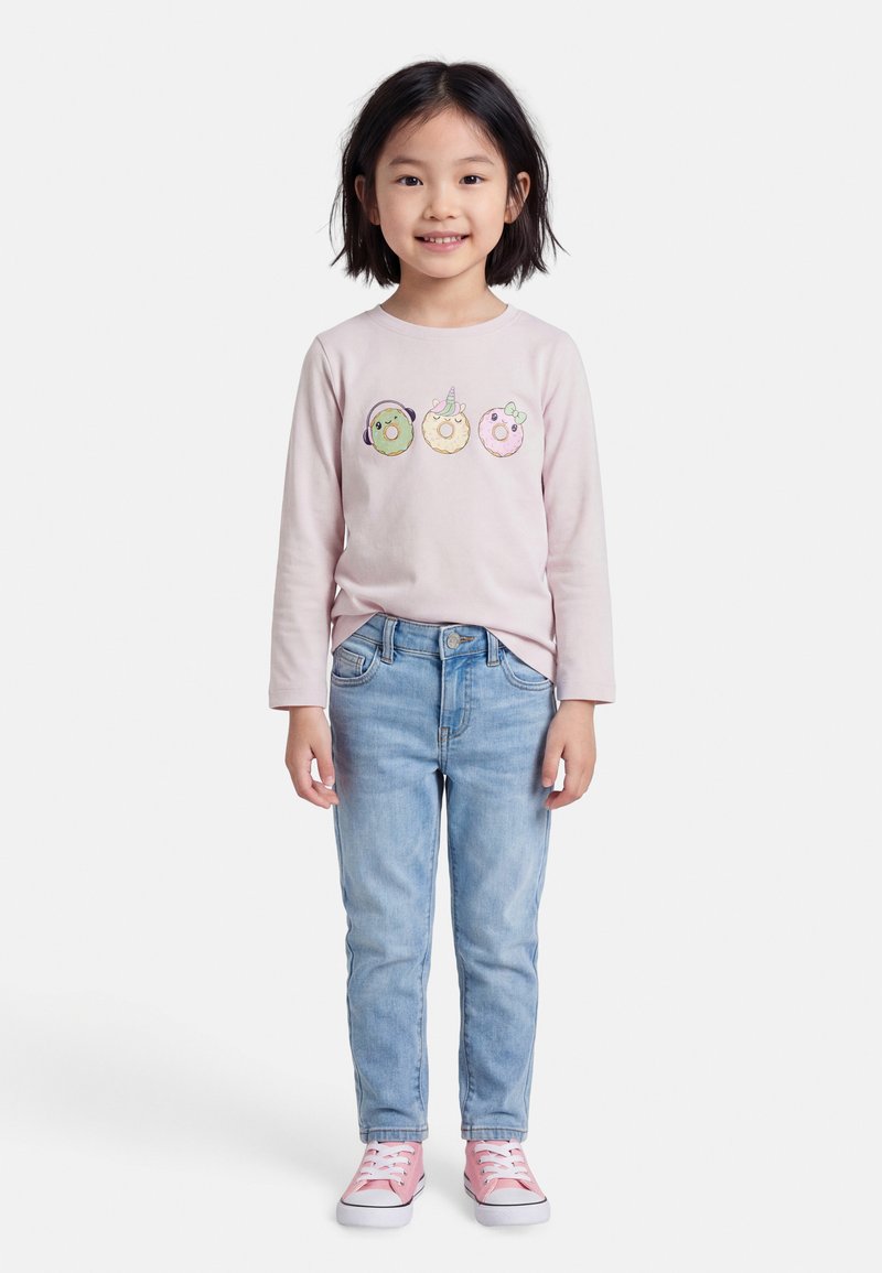 Ένα παιδί που χαμογελά φοράει ροζ μπλούζα με μακριά μανίκια και τρία παιχνιδιάρικα σχέδια ντόνατ, ανοιχτό μπλε τζιν και ροζ αθλητικά παπούτσια.