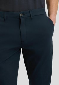Marineblaue Hosen mit glatter Textur, ausgestattet mit einem Knopfverschluss, Fronttaschen und einer taillierten Passform. Nahtdetails an der Vorderseite.