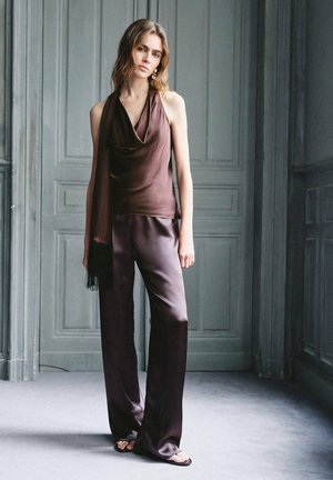 Mujer está de pie en una habitación gris vistiendo un top halter de satén marrón oscuro, pantalones anchos a juego, sandalias y pendientes dorados.