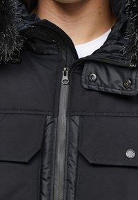 Schwarze gesteppte Jacke mit Reißverschluss, zwei großen Fronttaschen und abnehmbarem Kapuzen mit Kunstpelzbesatz. Sichtbare strukturierte Stoffdetails.