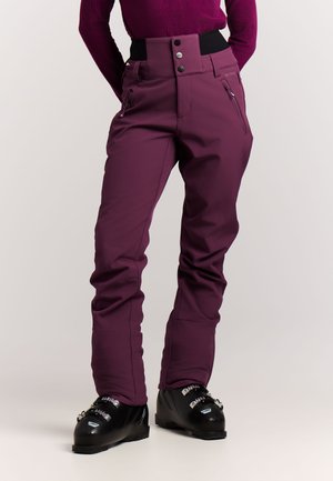 Persoon die paarse skisbroek met hoge taille, zwarte skischoenen met gespen en een paarse top met lange mouwen draagt tegen een effen achtergrond.
