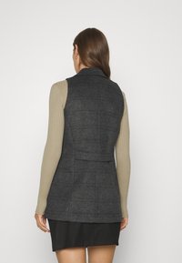 Gilet grigio a quadri con un design strutturato, caratterizzato da un colletto a notched e un pannello posteriore, indossato sopra una maglietta beige a maniche lunghe con costine.