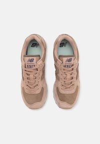 Ett par beige och bruna New Balance 574-sneakers sedd uppifrån mot en vit bakgrund.