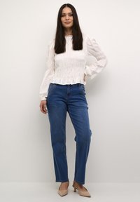 Blusa bianca texturizzata con maniche lunghe e bordo arricciato abbinata a jeans blu a gamba dritta e scarpe con tacco beige. Estetica semplice e moderna.