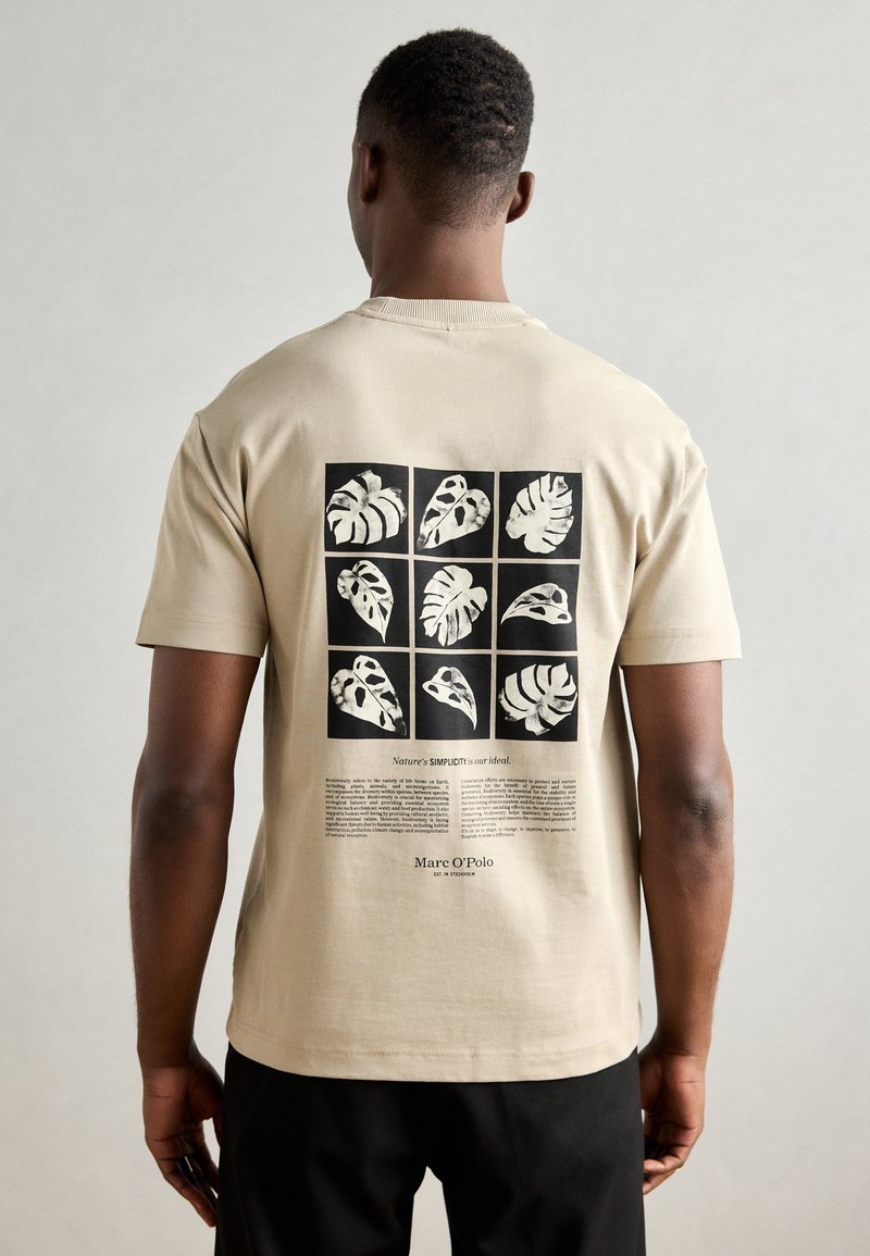 Beige cotton t-shirt featuring a black graphic with nine leaf prints and text. Short sleeves, crew neck, relaxed fit.