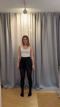 Noor naine valge varrukateta topi, mustade lühikeste pükste ja mustade sukkpükstega, seisab paljajalu puidust põrandal heledate hallide kardinate ees.