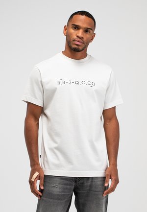 Mann trägt weißes T-Shirt mit schwarzen gedruckten Buchstaben und Pfeilen, kombiniert mit grauer Jeans, vor einfarbig hellem Hintergrund.