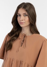 Lysebrun bluse lavet af blødt stof, med rund hals og snorelukning, samt puffermer og en lagdelt design ved kanten.
