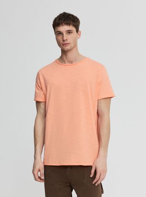 Giovane uomo che indossa una maglietta arancione chiaro a maniche corte semplice e pantaloni marroni, in piedi davanti a uno sfondo grigio chiaro uniforme.