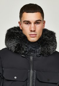 Svart pufferjakke med stor faux fur-krage. Har glidelås foran, to lommer foran og en glatt tekstur.