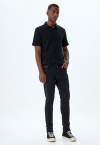 Camisa polo negra con una tela texturizada, combinada con jeans oscuros de corte ajustado. Footwear presenta un upper negro y una suela crema.
