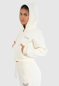 Crème-kleurige cropped hoodie gemaakt van zachte stof, met een voorzak, een koord en een geborduurd logo op de linkerborst.