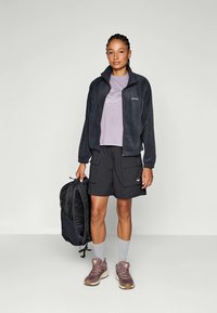 Giacca di pile grigia, t-shirt viola, pantaloni cargo neri, calze corte grigie e scarpe da ginnastica marroni; tiene uno zaino nero. Design minimalista.