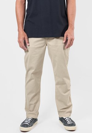 Homme portant un pantalon beige, des baskets noires avec des lacets blancs et une chemise bleu foncé, debout devant un fond uni.
