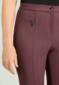Pantalones a medida color burdeos con una textura suave, que incluyen un bolsillo lateral con cremallera y una cinturilla ajustada. Detalle sutil de costura diagonal.