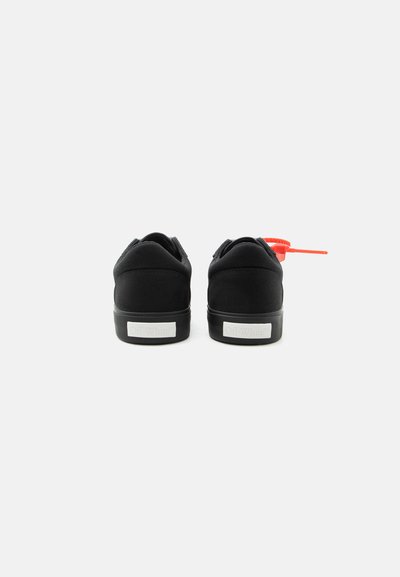 Paire de baskets basses noires vues de dos avec des étiquettes de talon blanches « Off-White » et une étiquette rouge à zip attachée à la chaussure droite.
