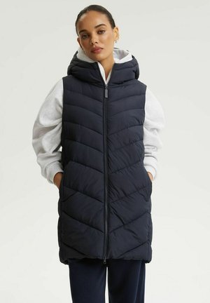 Kobieta ubrana w czarną pikowaną kamizelkę z kapturem na jasnoszarej bluzie z kapturem, z rękami w kieszeniach kamizelki, stoi na jednolitym tle.