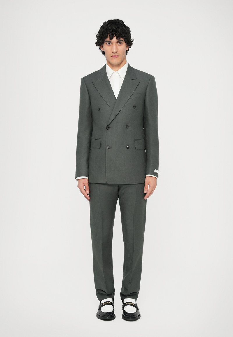 Abito doppiopetto verde scuro con lapels a punta e pantaloni abbinati, indossato con una camicia bianca e scarpe bianco e nere.