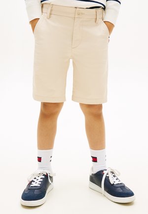 Bambino con pantaloncini beige, calzini bianchi con righe rosse e blu, e scarpe da ginnastica blu navy con lacci bianchi, in piedi con le mani nelle tasche.