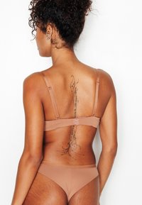 Set di lingerie marrone in tessuto morbido e liscio. Il reggiseno è con spalline regolabili e la modella ha un tatuaggio lungo la colonna vertebrale.