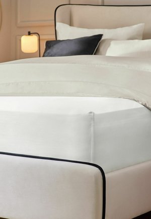 Bett mit beige gepolstertem Rahmen, weißem Spannbettlaken, cremefarbiger Bettdecke, zwei weißen Kissen, einem dunkelgrauen Kissen und einer eingeschalteten Nachttischlampe.