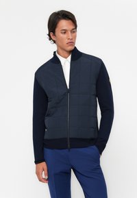Chaqueta acolchada navy con cuello alto y cremallera completa, que presenta un diseño ajustado de punto en las mangas y el dobladillo inferior, con una textura de tela suave.