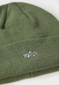 Căciulă tricotată verde cu o textură moale și un accent din metal argintiu. Prezintă un design aderent și un model cu nervuri.