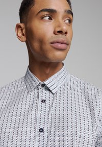 Overhemd met knopen gemaakt van een stof met een patroon van kleine ankers in het blauw op een lichte achtergrond. Bevat een kraag en donkere knopen.