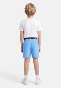 Παιδί που στέκεται με την πλάτη στην κάμερα φοράει άσπρο μπλουζάκι, ανοιχτό μπλε σορτς με πίσω τσέπη, άσπρες κάλτσες και άσπρα αθλητικά παπούτσια σε απλό φόντο.