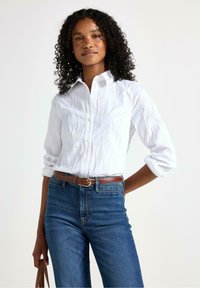 Hvid skjorte med knapper og tekstureret overflade samt opsmøgede ærmer, parret med højtaljede blå denimjeans og et brunt bælte.