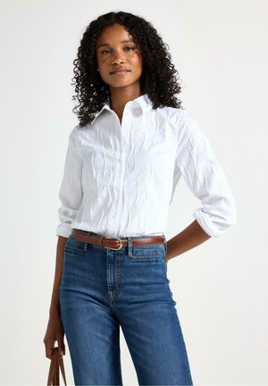 Camicia bianca con finitura texturizzata e maniche arrotolate, abbinata a jeans in denim blu a vita alta e una cintura marrone.