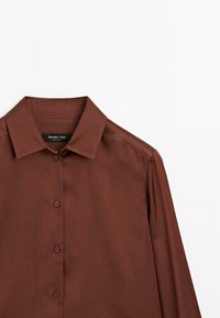 Chemise brune à manches longues en tissu lisse. Présente un col classique et une fermeture à boutons avec un design structuré subtil. Étiquette visible.