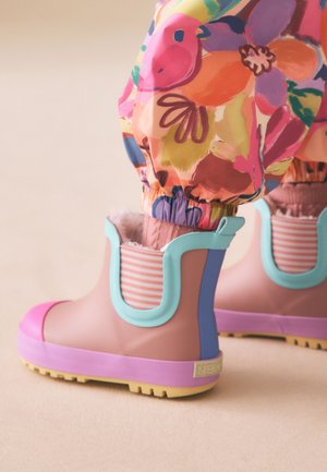 Gyermek, aki színes virágmintás nadrágot visel, ami pasztell gumicsizmába van tűrve; a csizmák rózsaszín orrnyereggel, lila talppal és akvamarin kék szegéllyel rendelkeznek, világos háttéren.